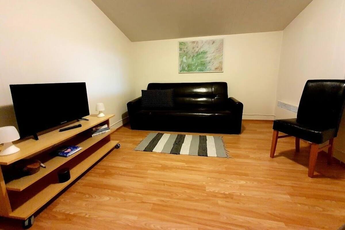 A living room with a black leather sofa, a wooden entertainment unit with a flat-screen TV, a remote control, and a lamp.