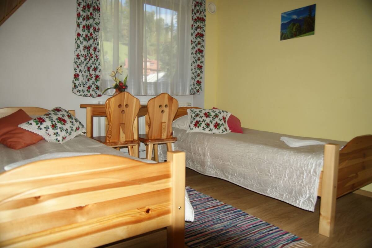 A bedroom with two wooden beds, a wooden chair, a small table, and a window with floral curtains.