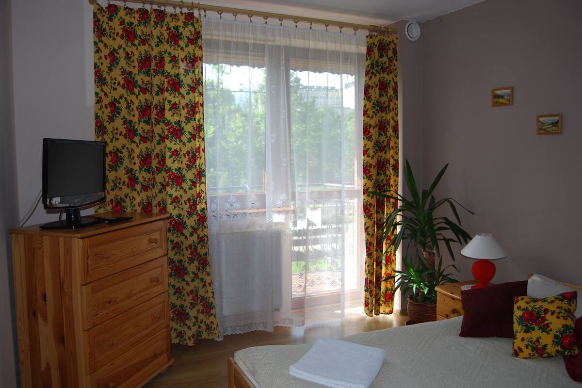 A bedroom with a wooden dresser, a bed with a white comforter, a television on the dresser, a potted plant, and a red lamp on a nightstand.
