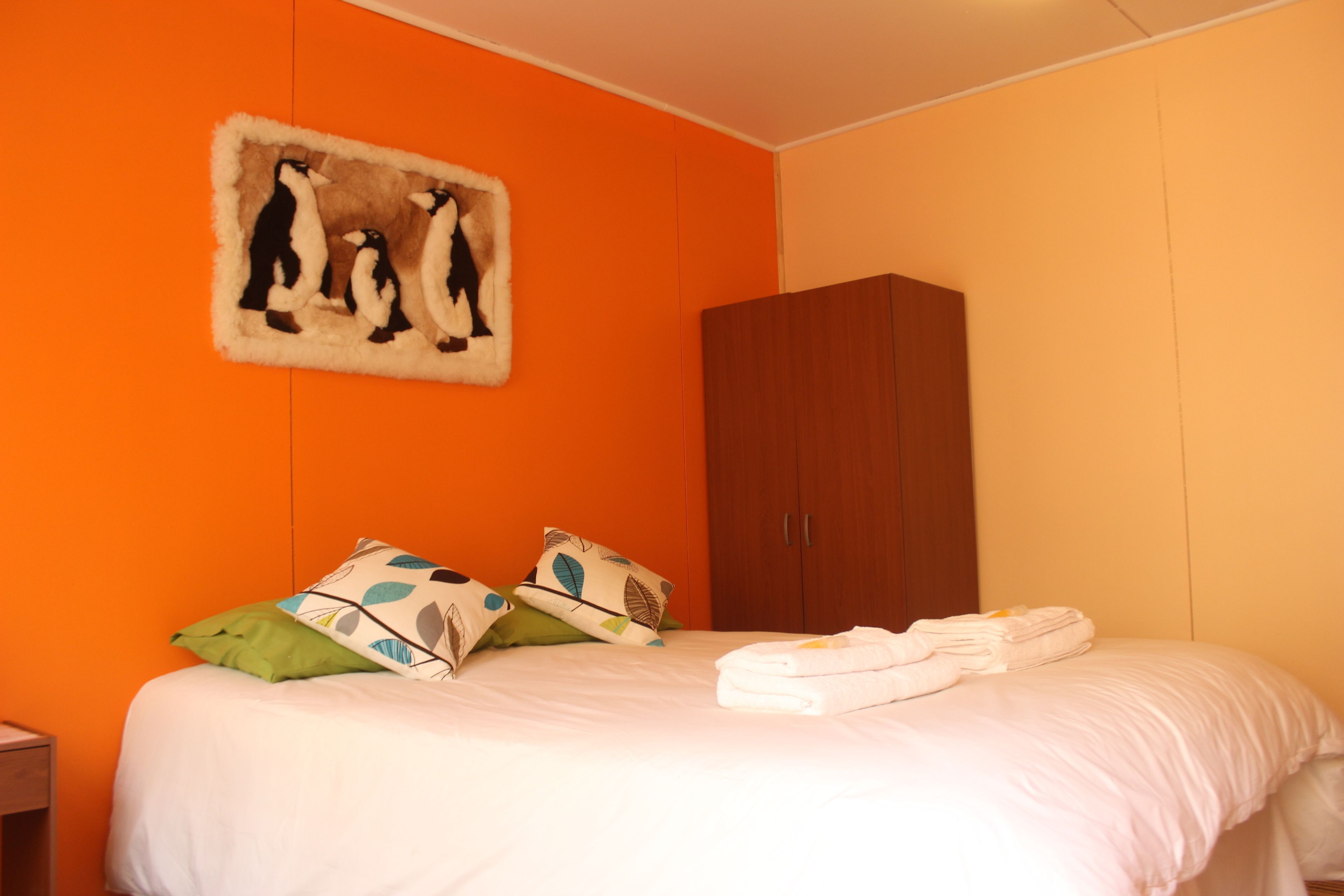 A bedroom with an orange wall, a bed with white bedding, a wooden wardrobe, and a wall art of penguins.