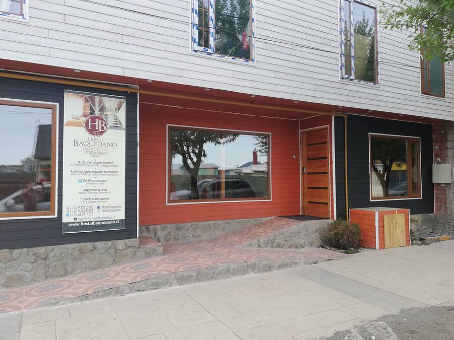 A building with a sign advertising a hotel named 'Hostal Baquedano' in Puerto Natales. It mentions 16 double rooms, a fully equipped private bathroom, Wi-Fi connection, a large dining area, and TV and board games.