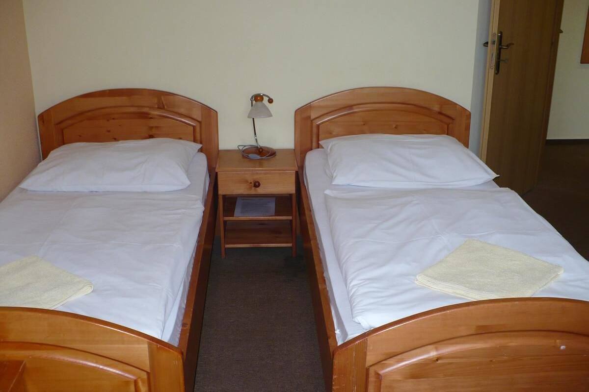 A room with two wooden beds, each with white linens and pillows. There is a small wooden nightstand between the beds with a lamp and a folded towel on it.