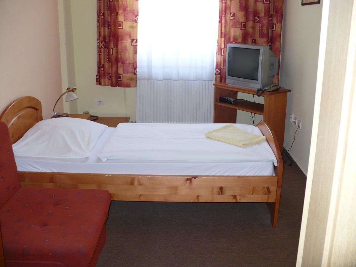 Ein Einzelbett mit Holzrahmen, ein kleiner Fernseher auf einem Holzständer, ein roter Sessel und ein Fenster mit gemusterten Vorhängen.
