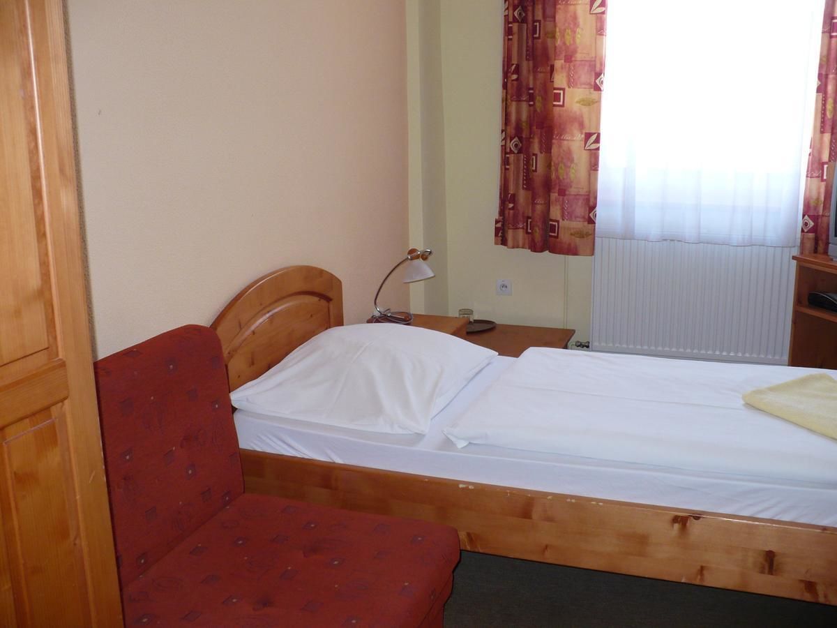 A single bed with a wooden headboard, a red upholstered bench, a bedside table with a lamp, and a window with patterned curtains.