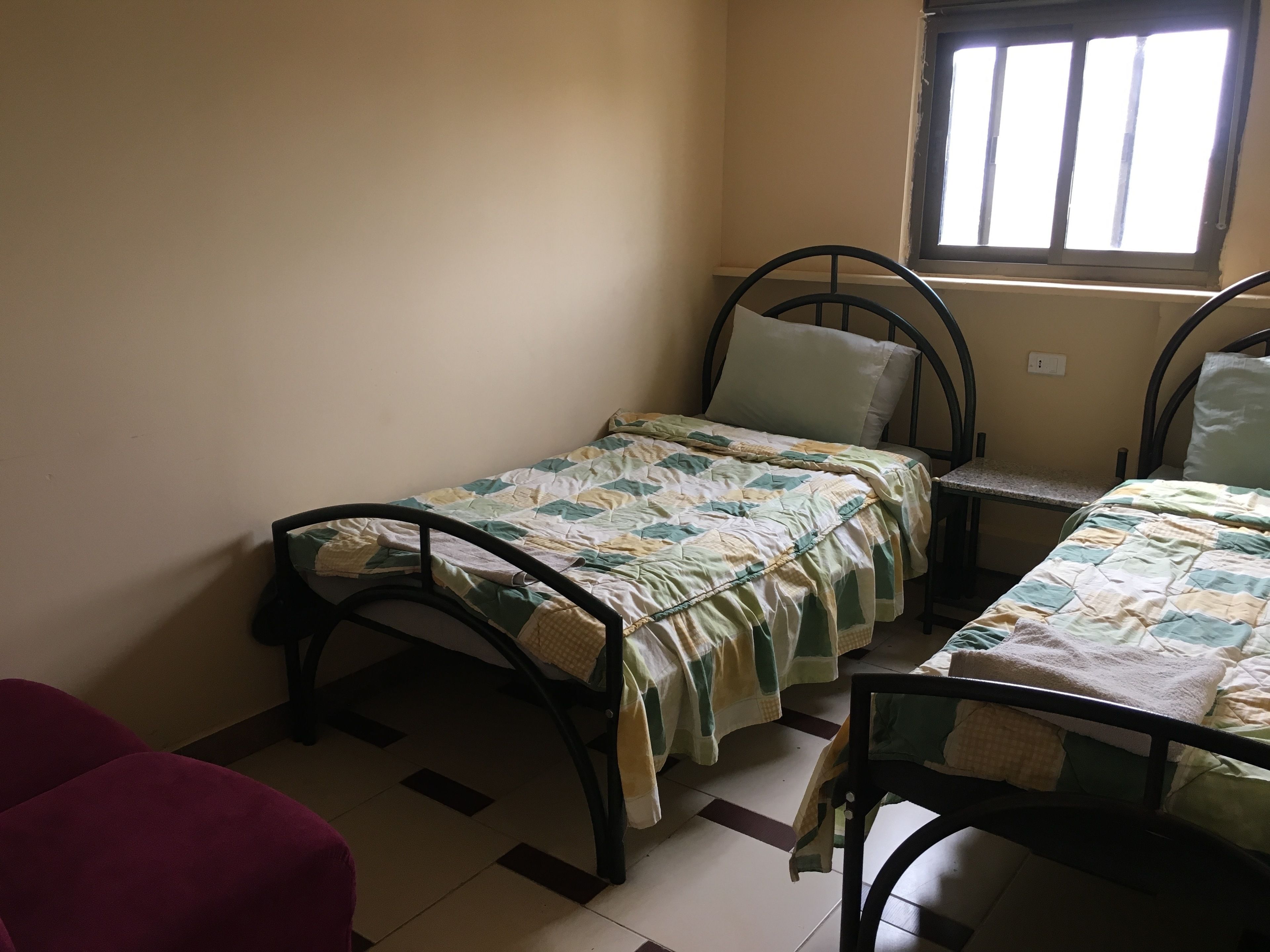 A small, single-room accommodation with two single beds, a red chair, and a window providing natural light.