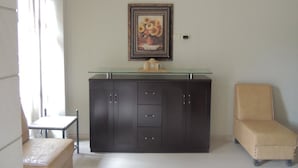 A room with a dark wooden cabinet, a glass-top table, a beige armchair, and a framed picture of flowers on the wall. - Madaba Hotel (Madaba)