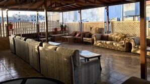 A covered outdoor seating area with sofas and a table, wooden beams on the ceiling, and a view of buildings in the background. - Madaba Hotel (Madaba)
