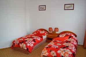 Een kamer met twee eenpersoonsbedden, elk met een bloemenprint deken, een houten nachtkastje met twee lampen en lijstjes met afbeeldingen aan de muur.