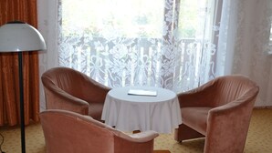 A room with two pink armchairs facing a small round table with a book on it. There is a floor lamp to the left and a window with a white lace curtain in the background. - Penzion Levandule (Brno)