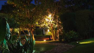 A garden pathway leading to a house with a well-lit porch area, surrounded by lush greenery.