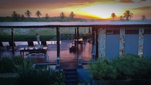 Uma área externa coberta com mesas, cadeiras, palmeiras e vista para o pôr do sol.