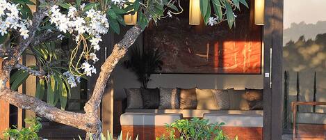 A modern outdoor seating area with a wooden bench and cushions, surrounded by lush greenery and flowering plants.