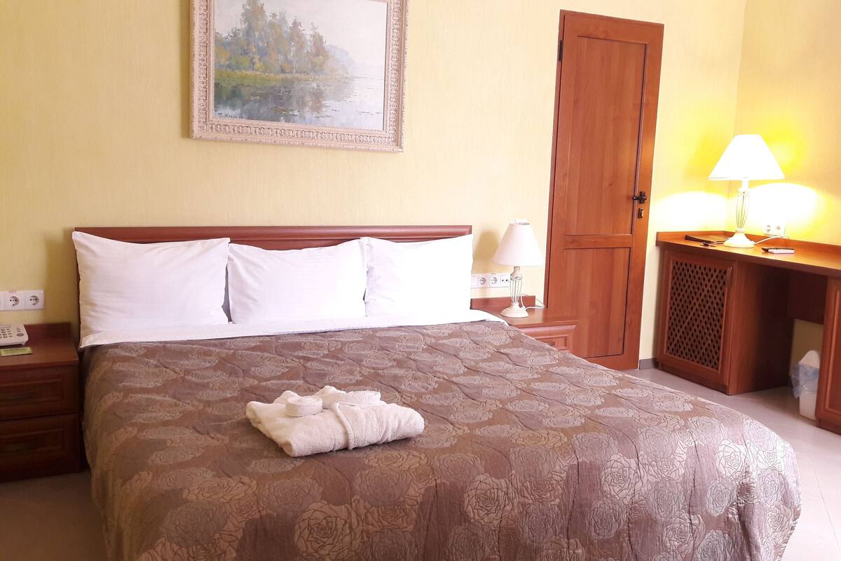 A hotel room with a large bed, a wooden nightstand, a desk with a lamp, a painting on the wall, and a door.