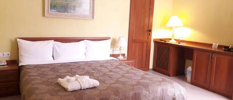 A hotel room with a large bed, a wooden nightstand, a desk with a lamp, a painting on the wall, and a door.