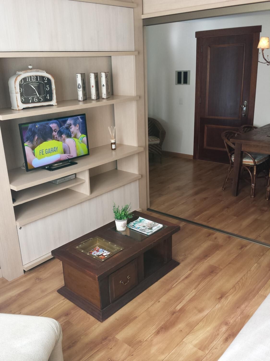 Uma sala de estar com piso de madeira, uma televisão sobre um suporte de madeira, uma mesa de centro com revistas e uma porta ao fundo.