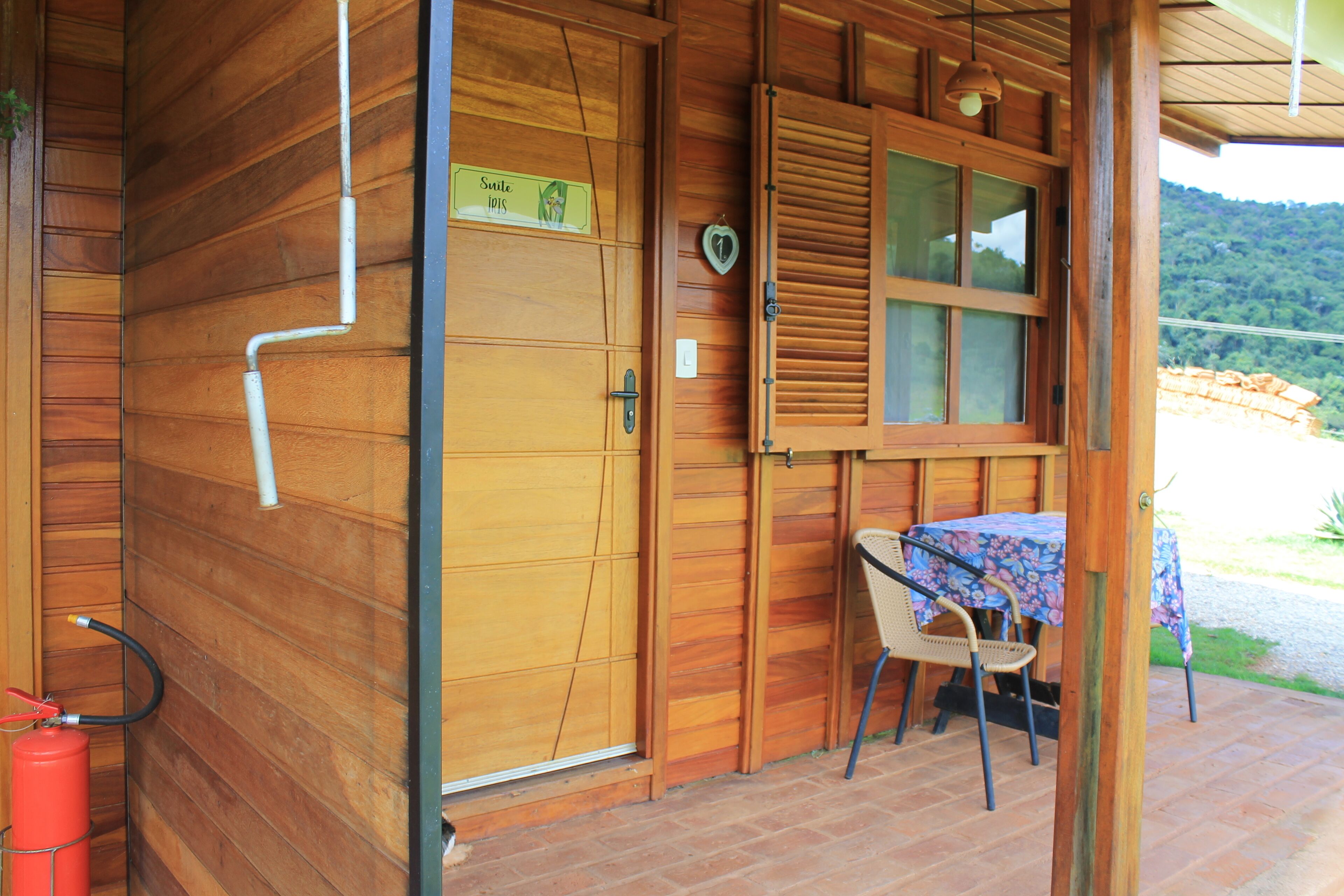 A wooden cabin with a closed door, a window with shutters, a table with a floral tablecloth, and a chair.