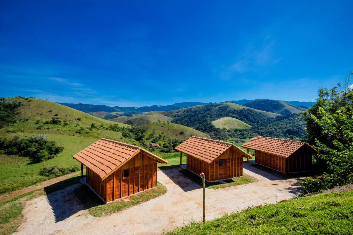 A rural setting with three wooden cabins with red roofs, surrounded by green hills and a clear blue sky.