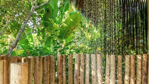 Una cerca de madera con listones verticales, un cabezal de ducha colgante y abundante follaje verde al fondo.