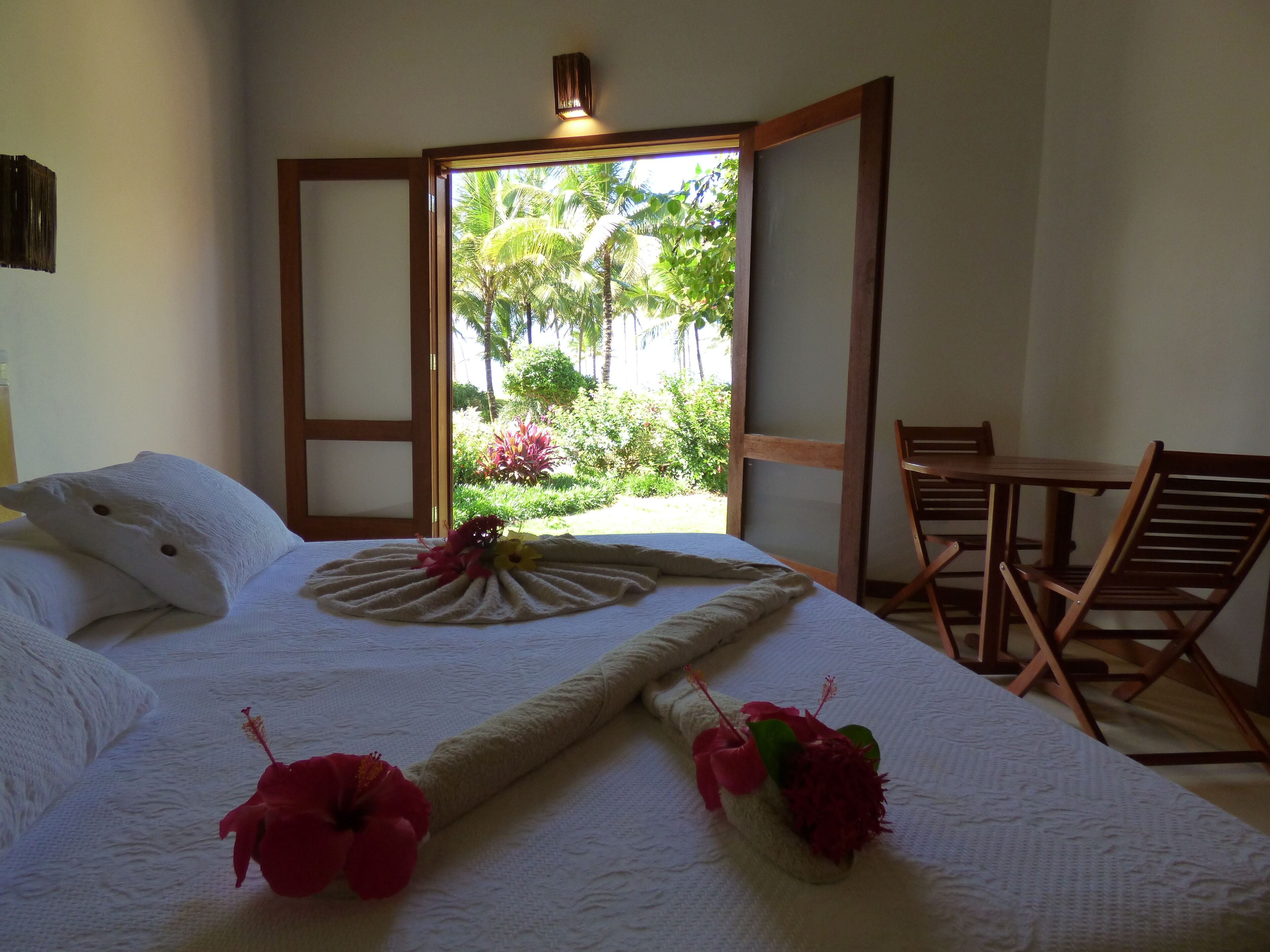 A neatly made bed with a decorative pillow and a floral arrangement, a wooden chair, and a view of greenery through an open door.