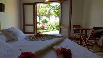 A neatly made bed with a decorative pillow and a floral arrangement, a wooden chair, and a view of greenery through an open door.