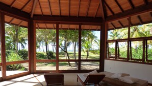 Habitación con techo de madera y ventanas amplias que dan a un paisaje tropical con palmeras y una playa. Hay dos sillas de madera y un banco con almohadones.