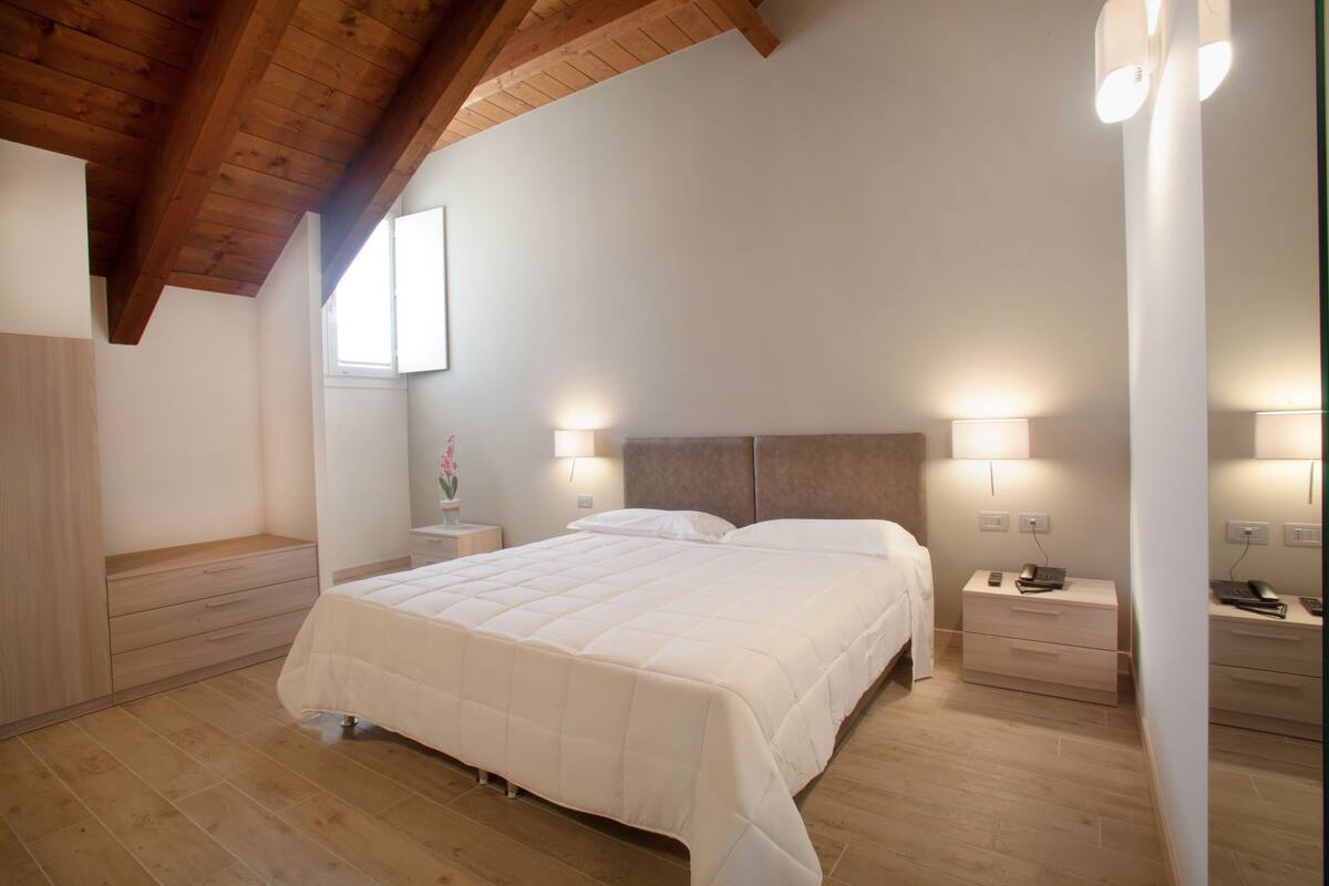 A bedroom with a wooden ceiling, a bed with a white duvet, bedside tables with lamps, and a window with natural light.