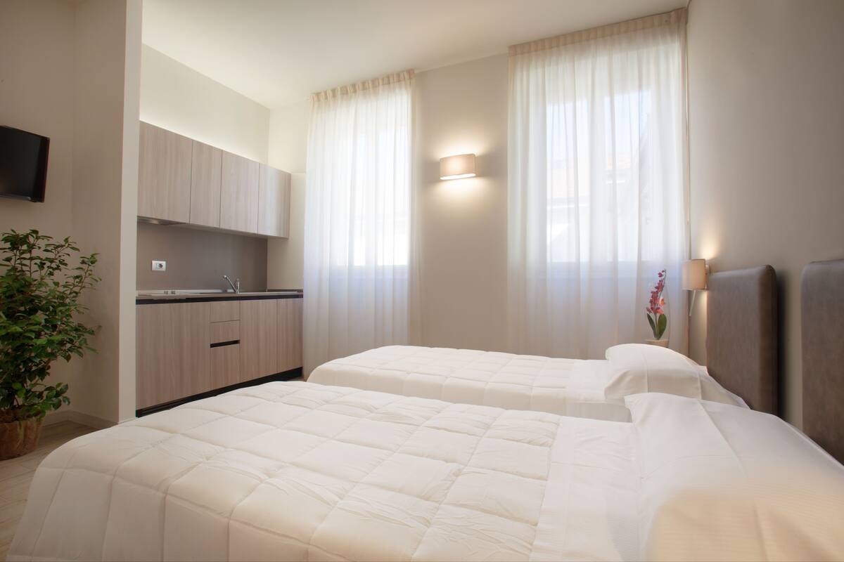 A hotel room with two beds, a kitchenette, a flat-screen TV, and a potted plant.