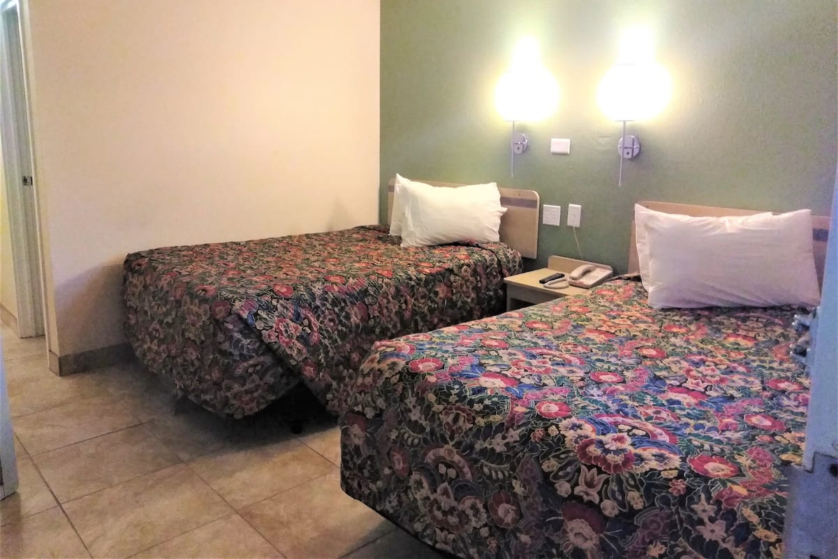 A hotel room with two beds, each with floral bedding, a small bedside table with a phone, and wall-mounted lights.