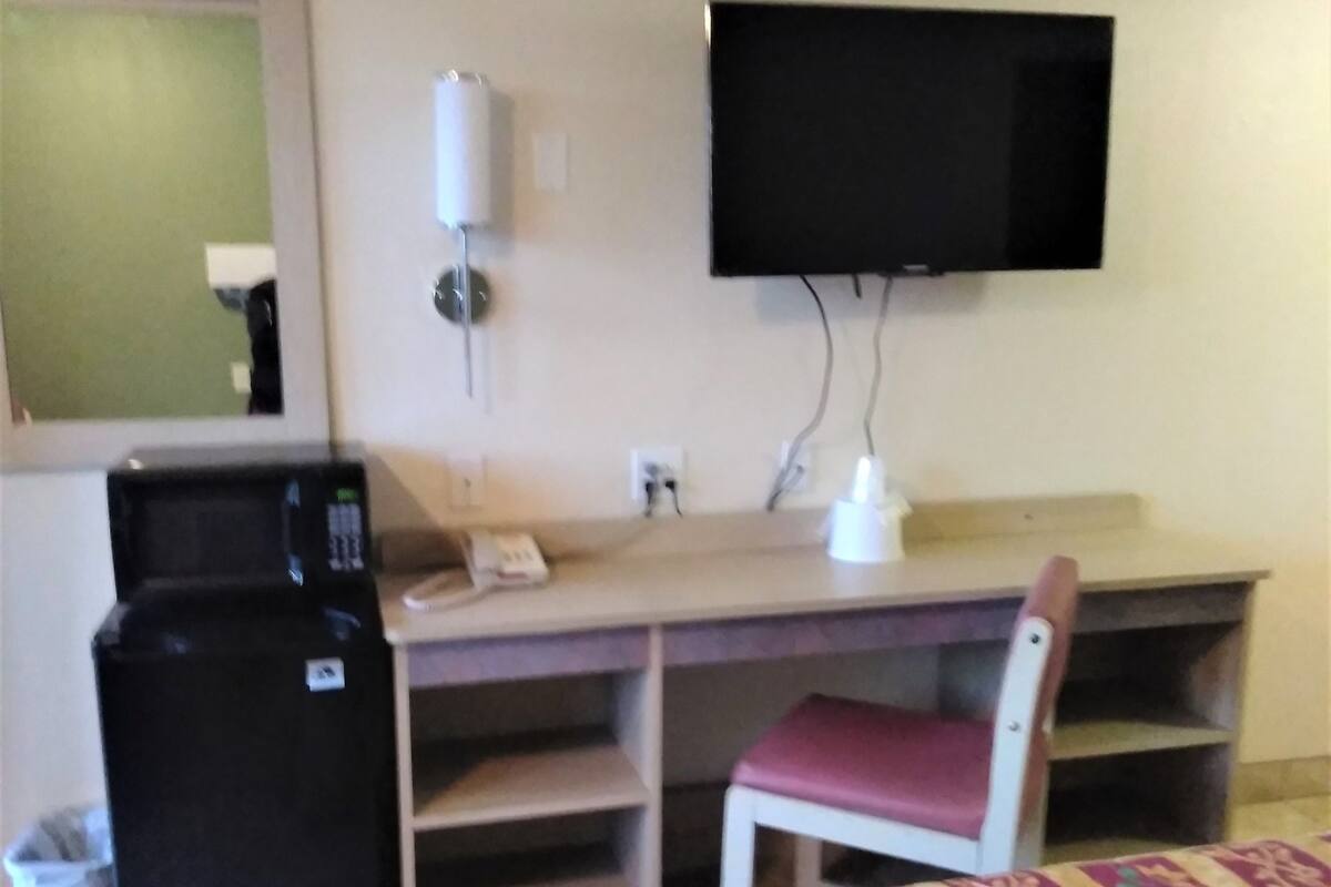 A hotel room with a bed, a desk with a chair, a television mounted on the wall, a refrigerator, and a trash bin.