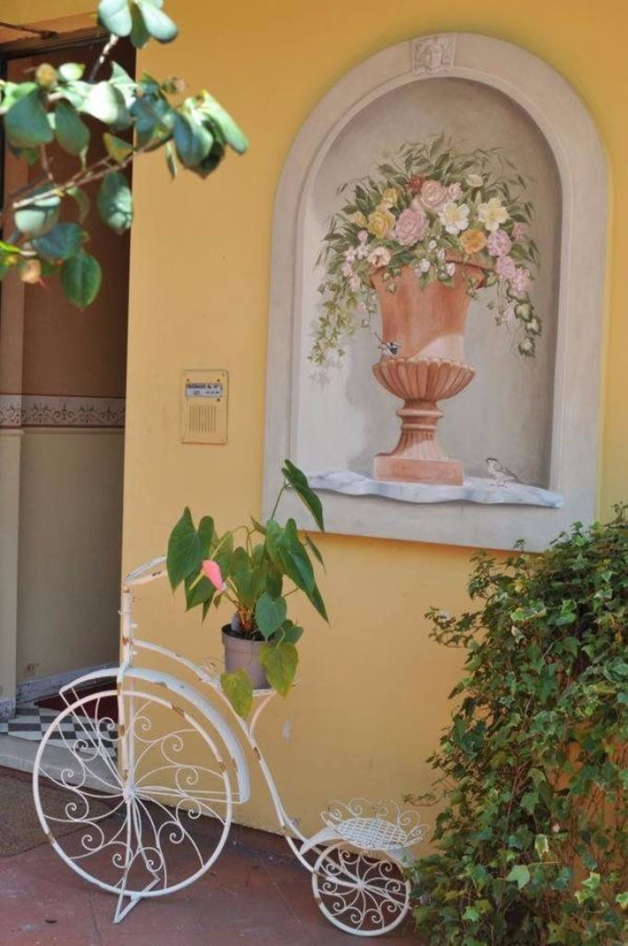 Un muro giallo con un murale raffigurante un vaso di fiori, una bicicletta in metallo bianca e una pianta in vaso.