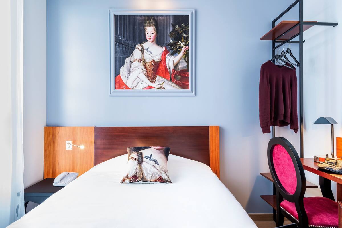 Ein Hotelzimmer mit Bett, Holz-Kopfseitenverkleidung, Nachttisch mit Telefon, gerahmtes Porträt an der Wand, Schreibtisch mit Lampe und Stuhl.