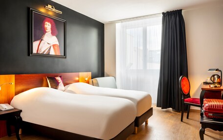 A hotel room with two beds, a desk with a telephone, a chair, a table with a lamp, a painting on the wall, and a window with curtains.