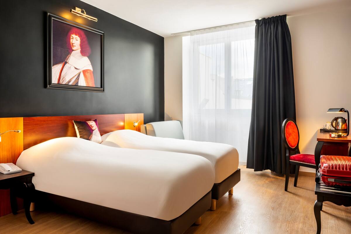 Ein Hotelzimmer mit zwei Betten, einem Schreibtisch mit Telefon, einem Sessel, einem Tisch mit Lampe, einem Bild an der Wand und einem Fenster mit Vorhängen.