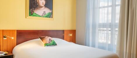 Una habitación de hotel con una cama grande, un cuadro en la pared, una ventana con cortinas y una mesita de noche con lámpara.