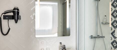 Un baño con lavamanos blanco, espejo, secador de pelo, ducha y una botella de champú.