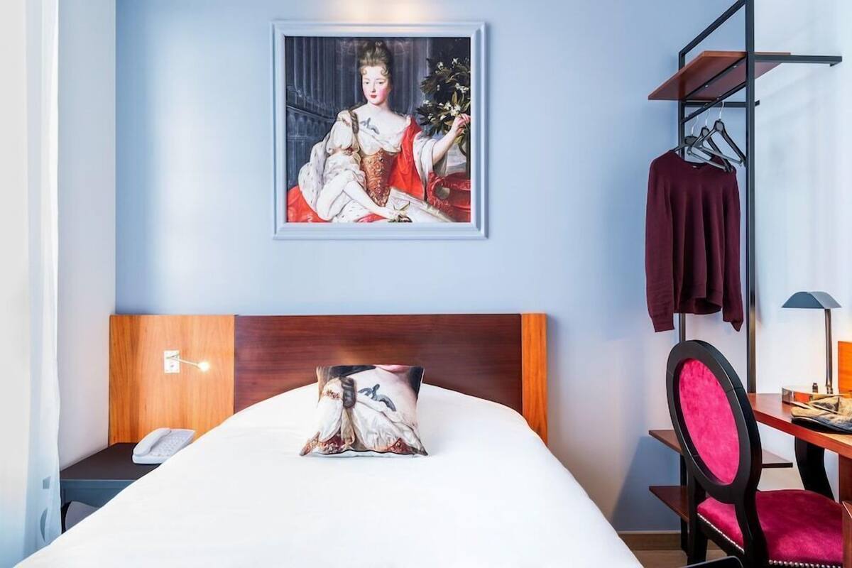 Ein Hotelzimmer mit Bett, Holz-Kopfteil, gerahmtem Gemälde an der Wand, Schreibtisch mit Stuhl und einer Garderobe.