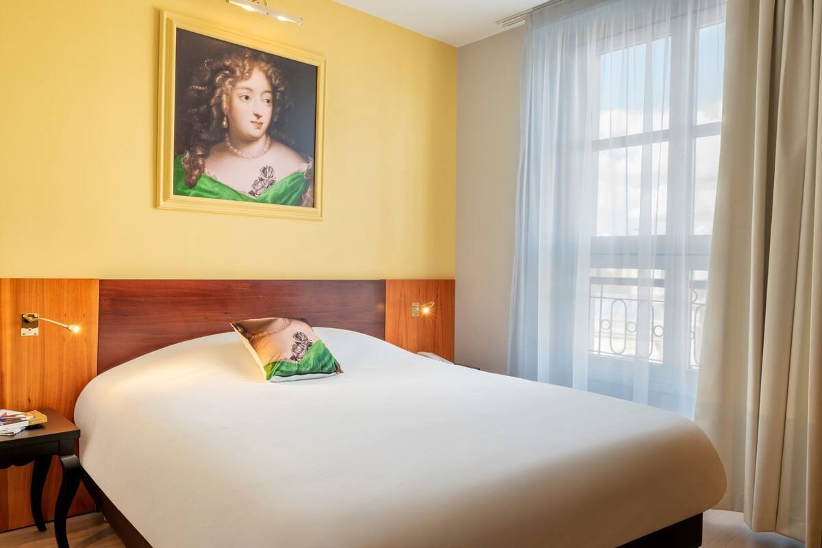 Ein Hotelzimmer mit einem großen Bett, einem Gemälde an der Wand, einem Nachttisch und einem Fenster mit Vorhängen.