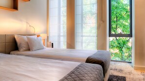 A hotel room with two beds, a large window with a view of greenery, and a wooden shelf above the beds.