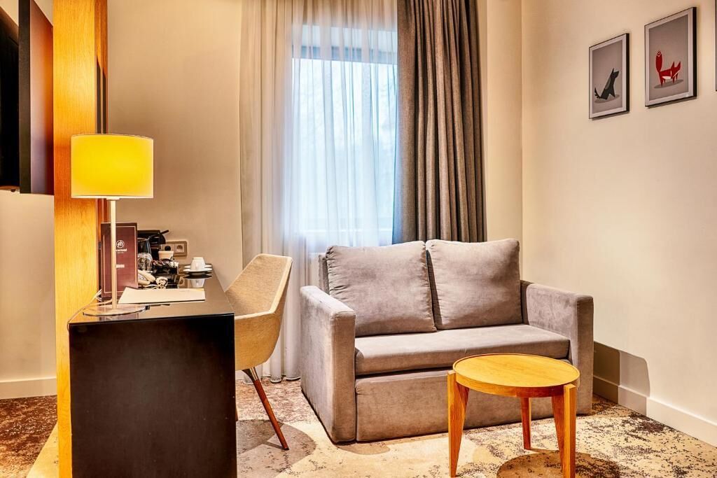 A modern hotel room with a grey sofa, a wooden side table, a desk with a lamp, and a window with sheer curtains.