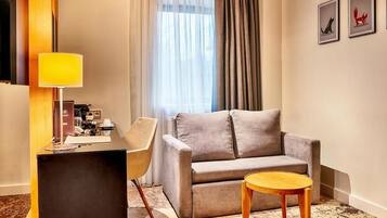 A modern hotel room with a grey sofa, a wooden side table, a desk with a lamp, and a window with sheer curtains.
