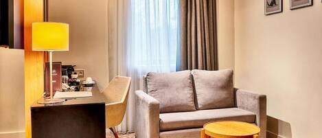 A modern hotel room with a grey sofa, a wooden side table, a desk with a lamp, and a window with sheer curtains.