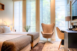 Una habitación de hotel con una cama, dos mesitas de noche, un escritorio con una silla, un televisor y ventanas amplias con vistas a árboles.