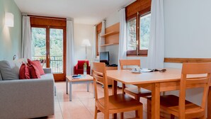Un salon moderne avec un canapé gris, une table à manger en bois et des chaises. Un téléviseur est fixé au mur, et on aperçoit un balcon à travers les fenêtres.