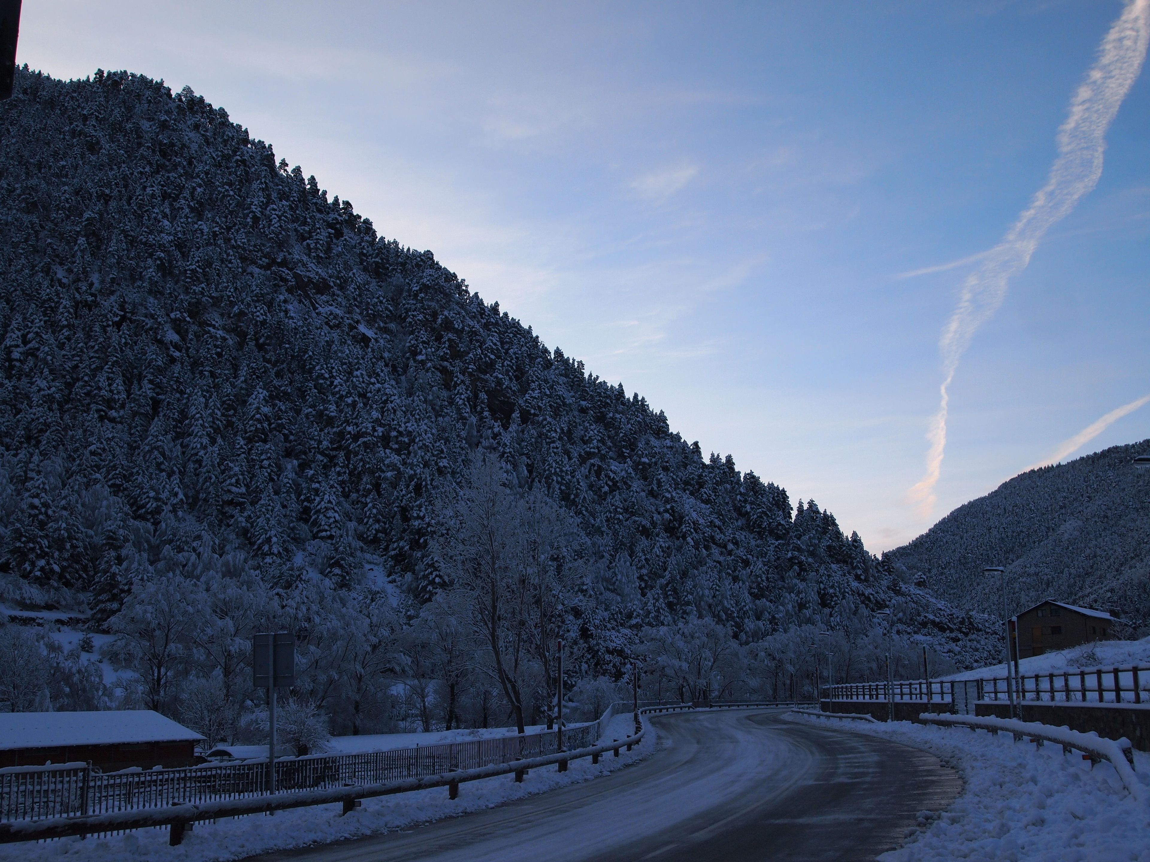 A snow-covered road curving through a hilly area with a wooden fence and a building on the right.