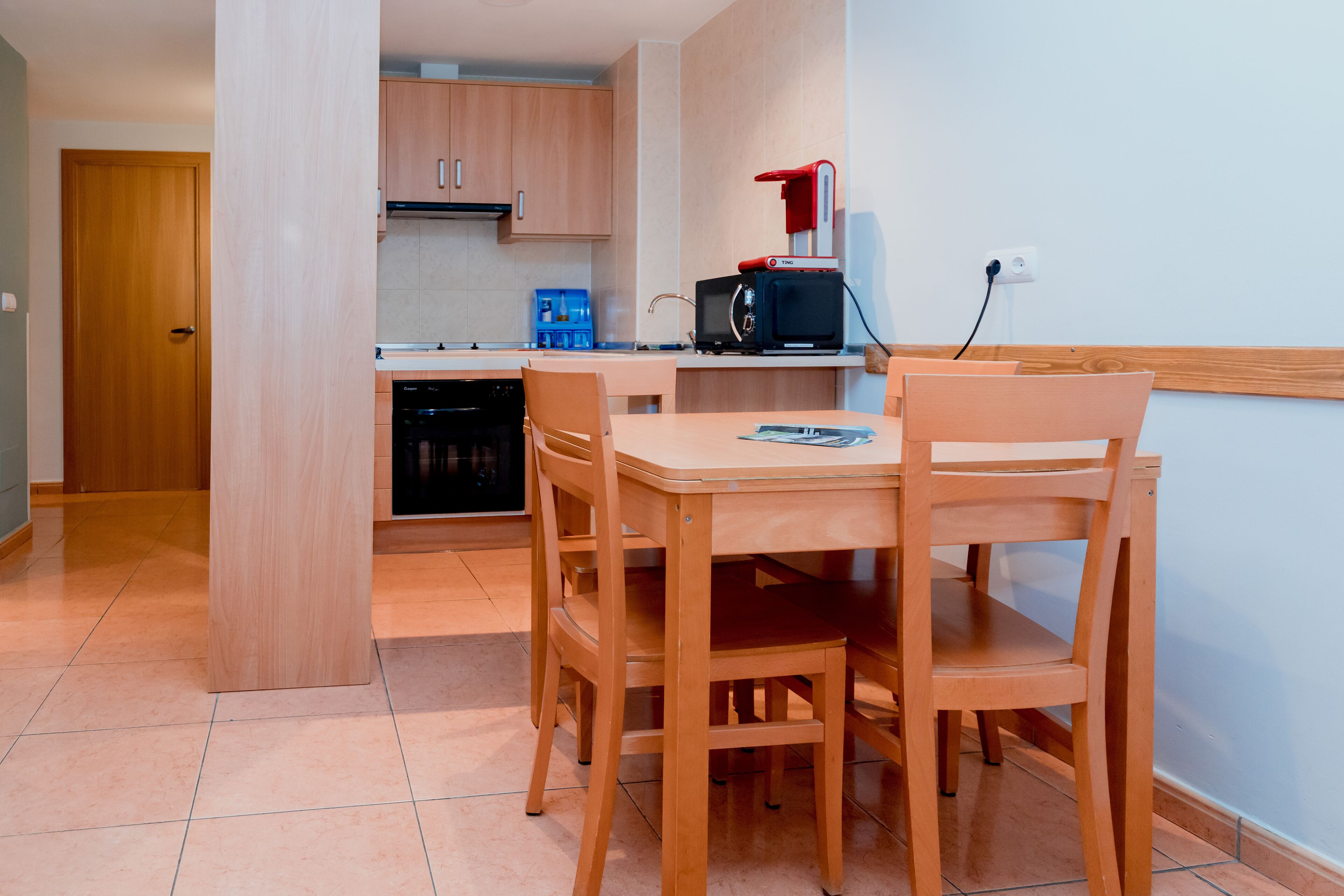 Een compacte keuken met houten kastjes, een magnetron en een kleine eettafel met stoelen.