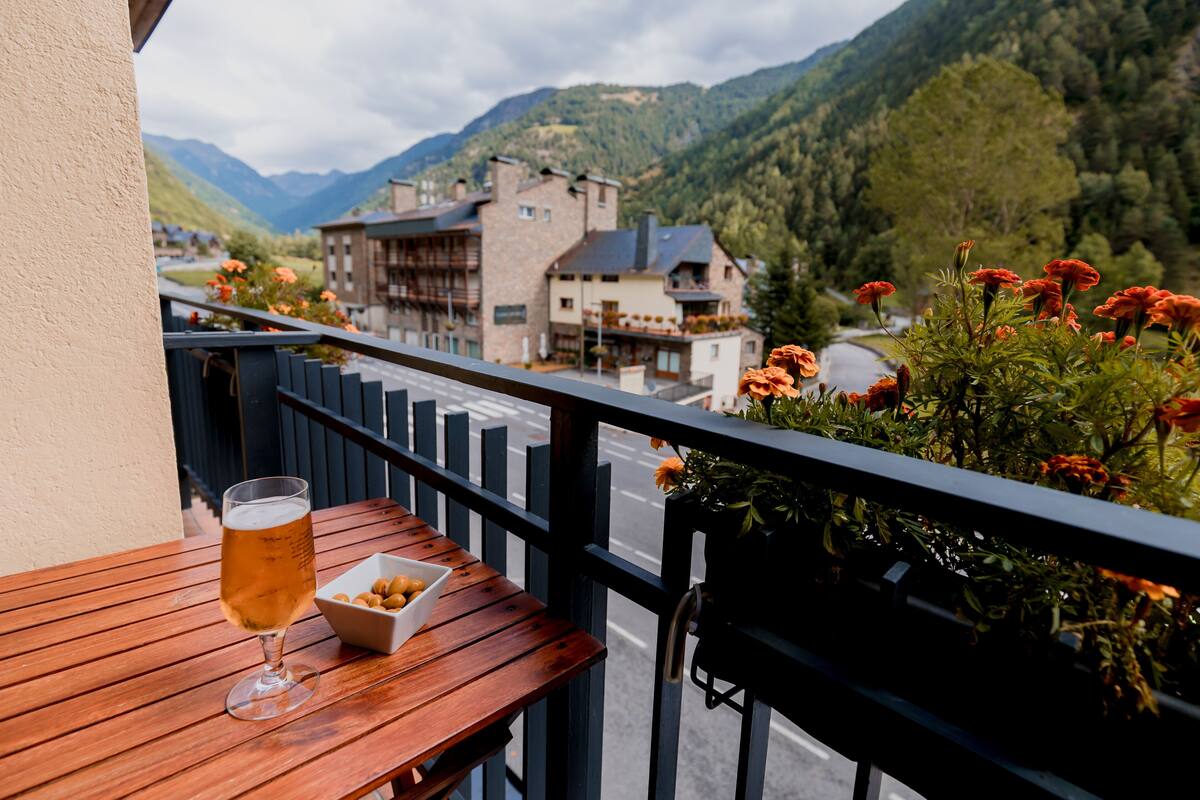 Een houten tafel met een glas bier en een schaal lekkernijen op een balkon met uitzicht op een berglandschap.