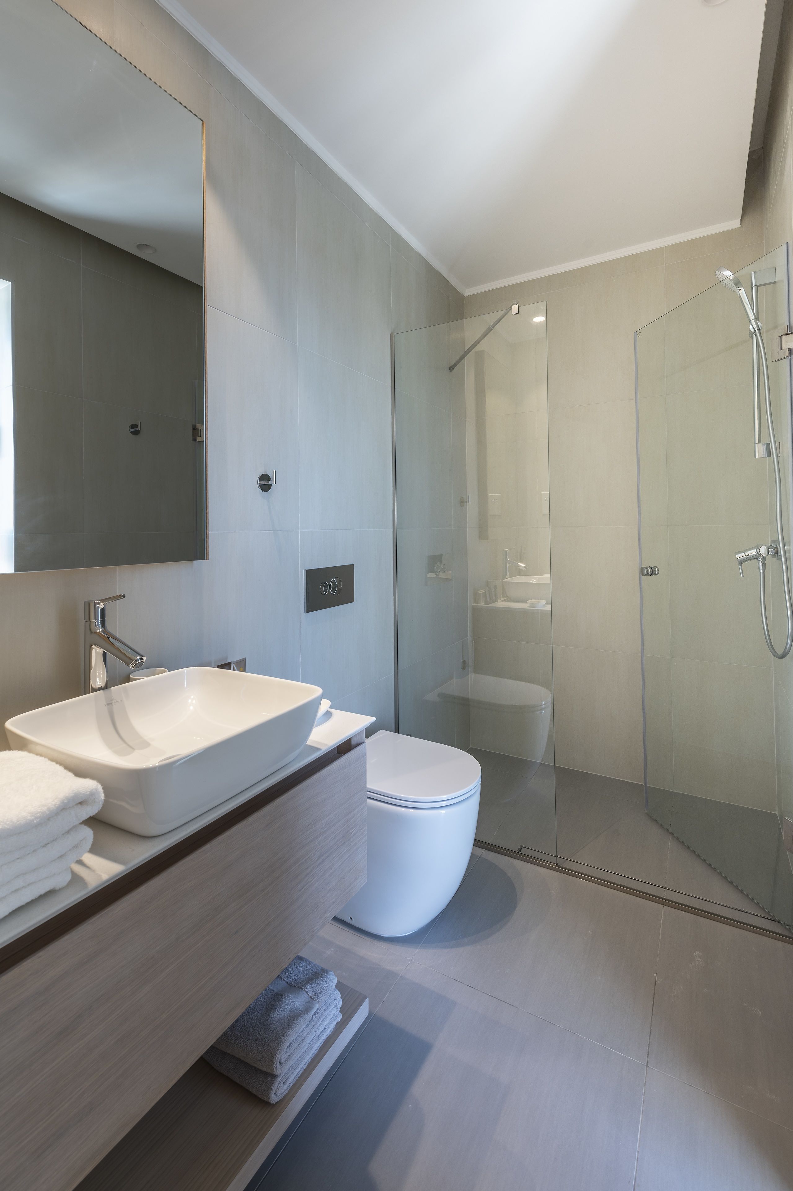 Un baño moderno con un espejo grande, un lavamanos blanco, un inodoro y una zona de ducha con mampara de vidrio.