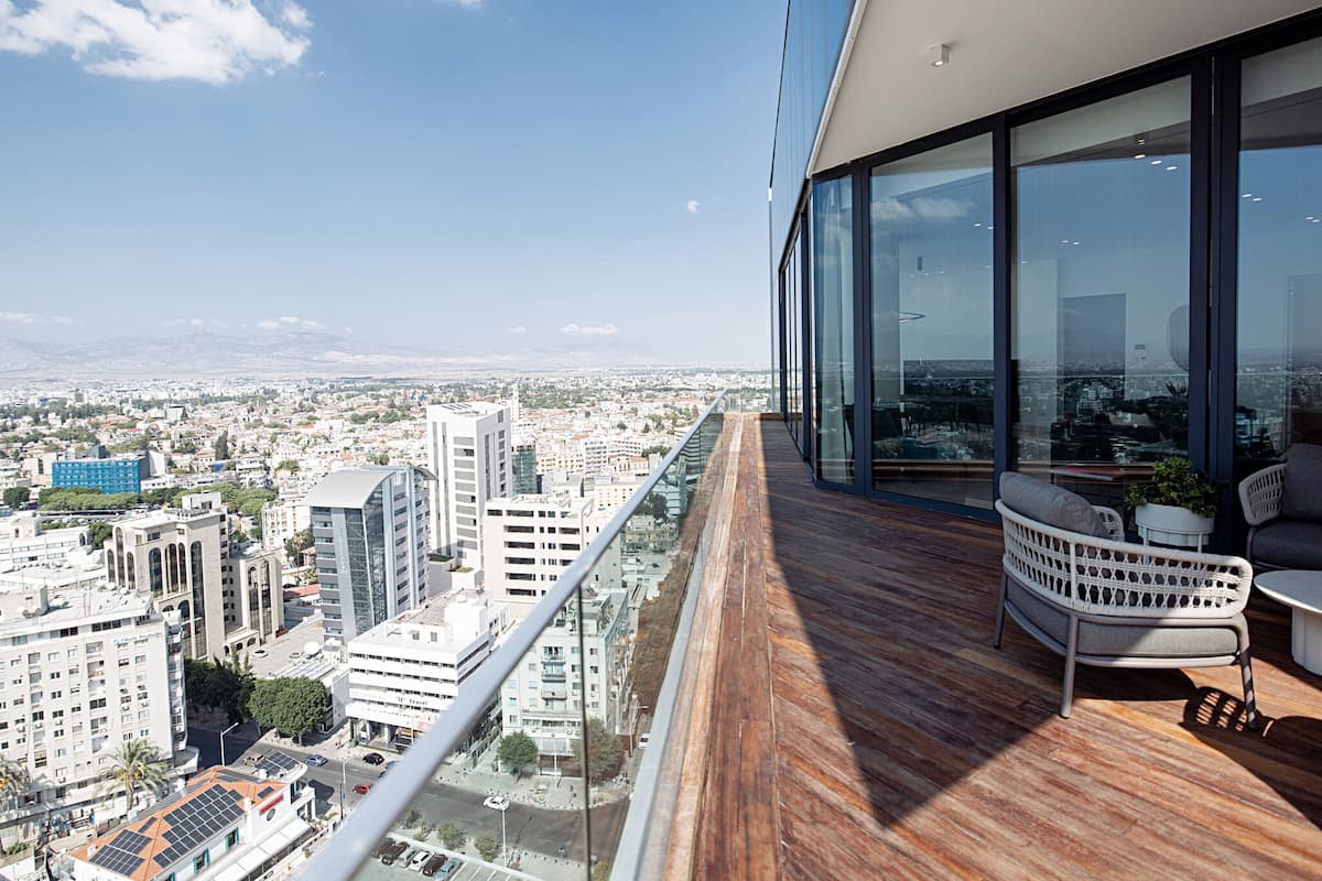Balcón de un edificio alto con vistas a la ciudad, suelo de madera y zona de estar con cojines y una pequeña mesa.