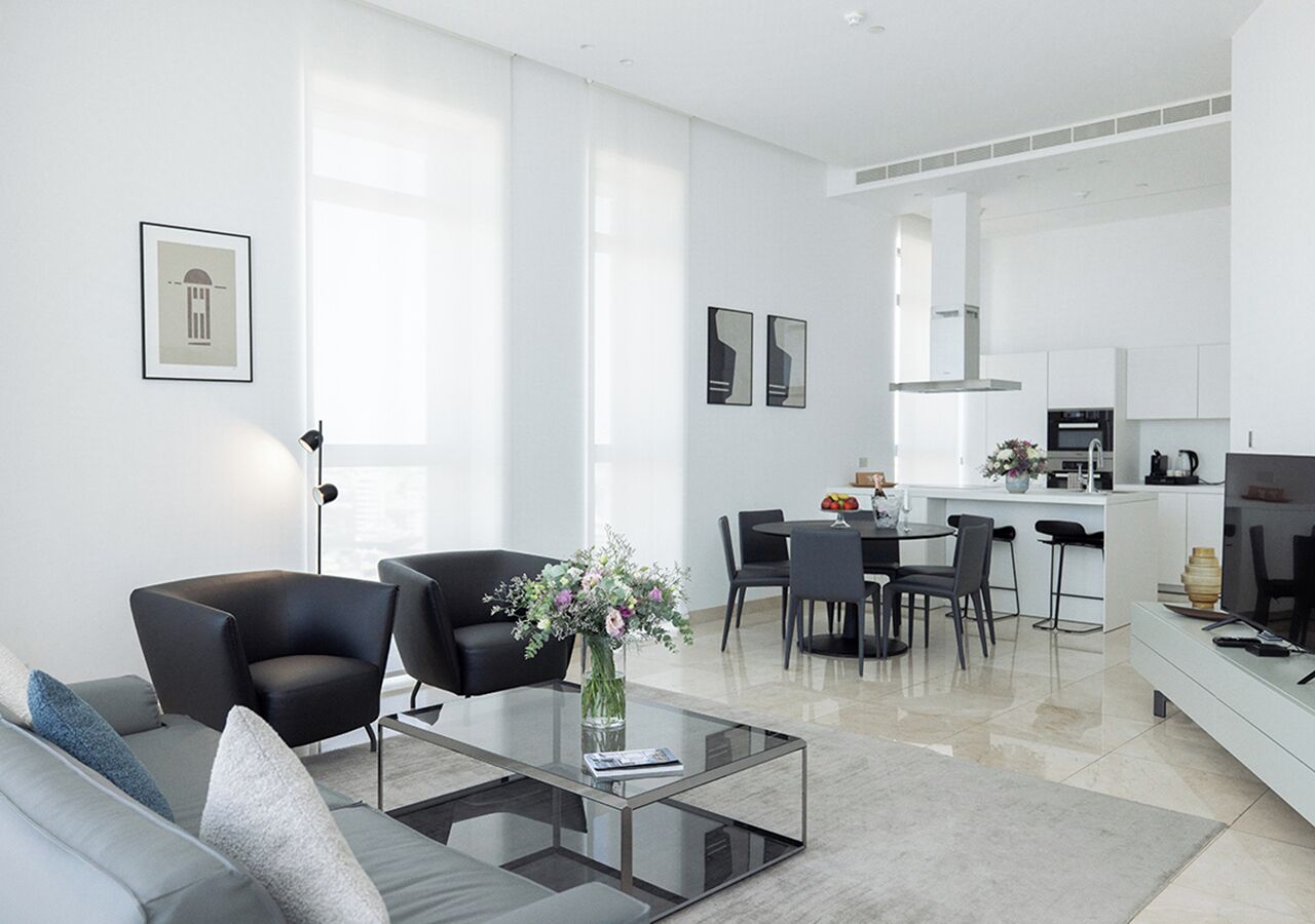 A modern living room with a glass coffee table, black armchairs, and a sofa. There is a dining area with a table and chairs, and a kitchen in the background.