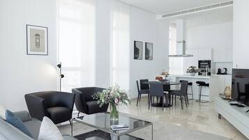 A modern living room with a glass coffee table, black armchairs, and a sofa. There is a dining area with a table and chairs, and a kitchen in the background.